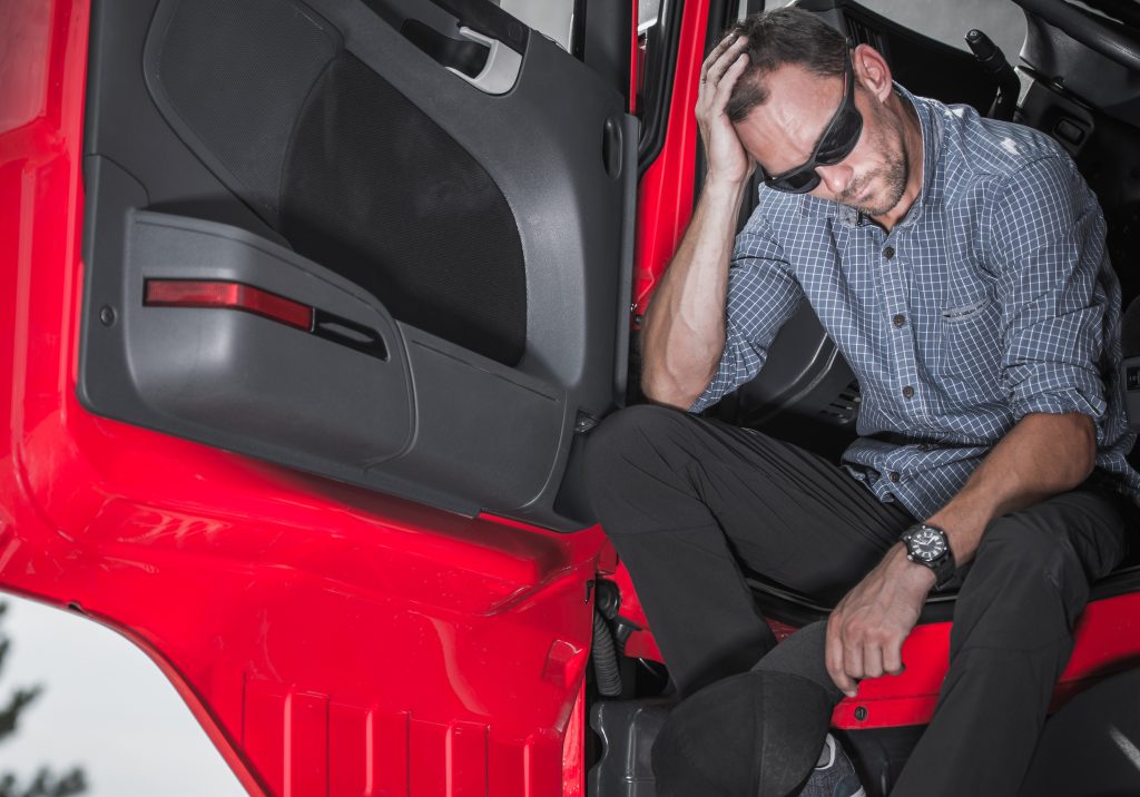 Un hombre angustiado, con gafas de sol y camisa a cuadros, sentado en el interior de un camión rojo, apoya la cabeza en la mano, lo que indica agotamiento o una situación estresante. Relevante para temas sobre el bienestar de los camioneros o la importancia de buscar asistencia legal tras un accidente de camión.