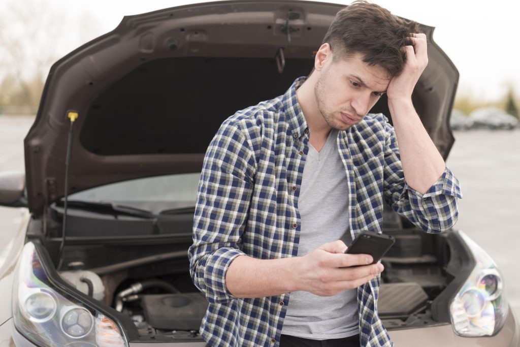 Un hombre estresado mirando su teléfono junto a un auto averiado, resaltando la importancia de comprender las 'Causas Comunes de Accidentes Automovilísticos en el Condado de Gwinnett'. Big Time Law está aquí para ayudar cuando necesite apoyo.